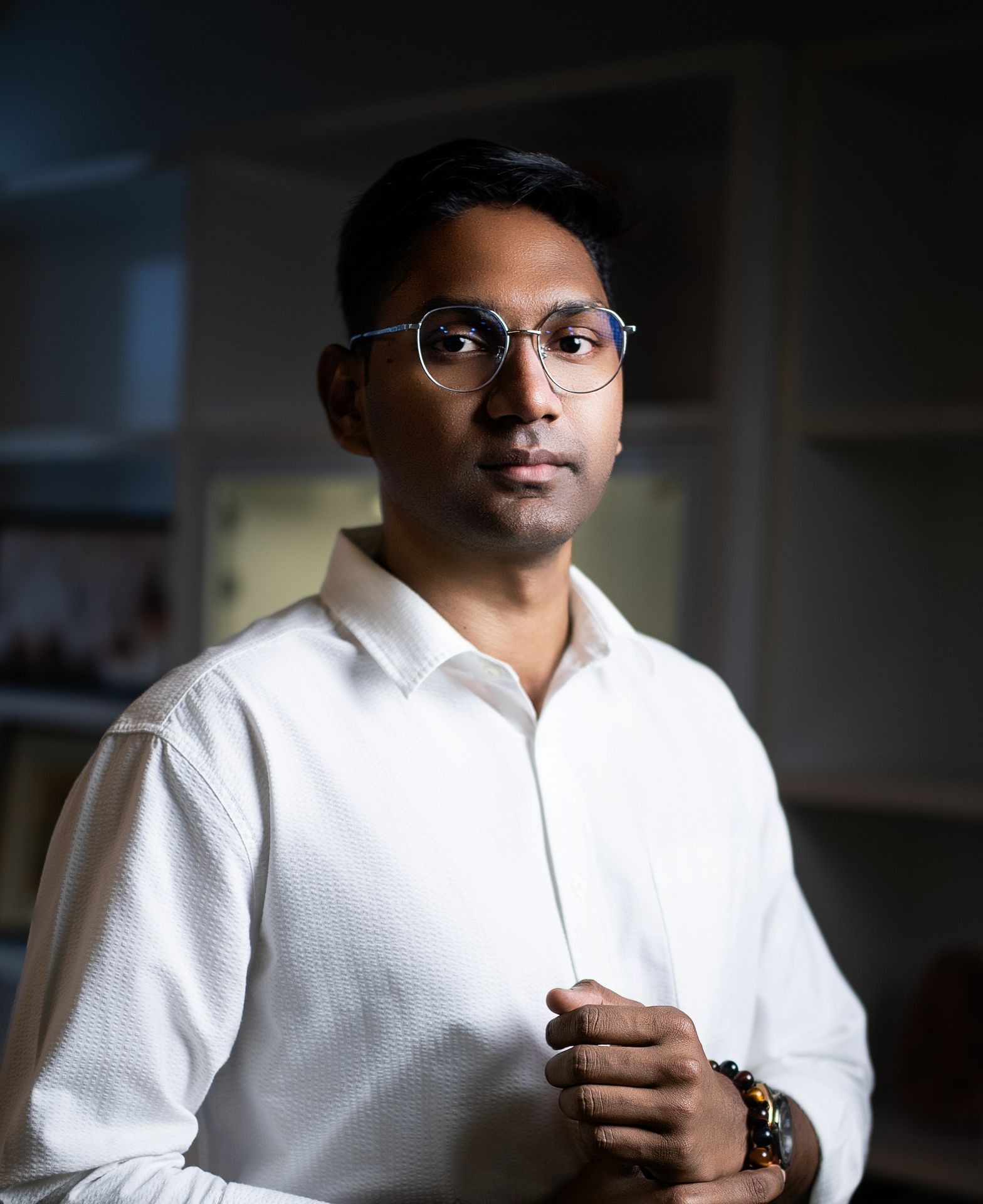 Person wearing glasses and a white shirt, standing in a softly lit room.